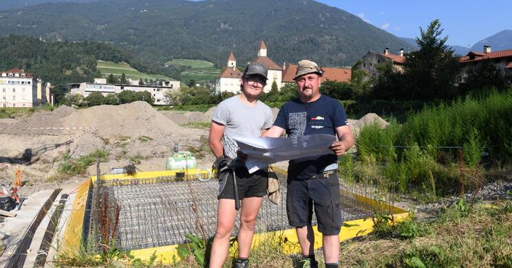 Zwei Arbeiter stehen auf der Baustelle im Hofburg Garten und blicken in die Kamera; im Hintergrund sind Gebäude und bewaldete Berge zu sehen.