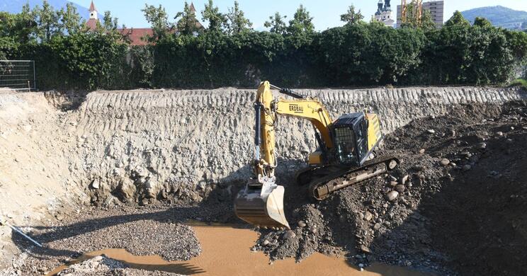 Ein Bagger gräbt in einem großen Erdloch auf der Baustelle des Hofburg Gartens; im Hintergrund Sträucher, die Türme des Doms und bewaldete Berge.