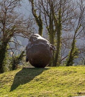 Eine große, abgerundete Steinskulptur mit Gesichtszügen und Ohren steht kopfüber auf einem grasbewachsenen Hügel; im Hintergrund kahle Bäume.