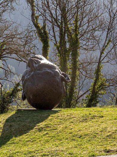 Eine große, abgerundete Steinskulptur mit Gesichtszügen und Ohren steht kopfüber auf einem grasbewachsenen Hügel; im Hintergrund kahle Bäume.