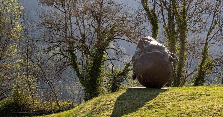 Eine große, abgerundete Steinskulptur mit Gesichtszügen und Ohren steht kopfüber auf einem grasbewachsenen Hügel; im Hintergrund kahle Bäume.