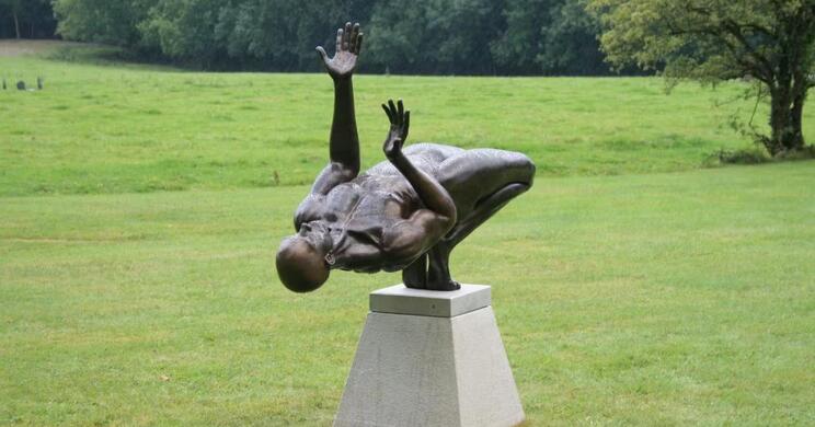 Eine dunkle, lebensgroße Statue eines nackten tanzenden Mannes auf einem hellen Sockel inmitten einer grünen Wiese; im Hintergrund sind Bäume zu sehen.