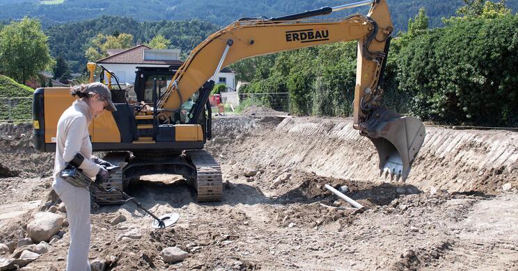 Ein Bagger auf der Baustelle im Hofburg Garten; daneben steht eine Arbeiterin, im Hintergrund sind bewaldete Berge zu sehen.