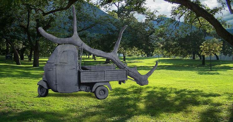 Eine graue Skulptur eines Piaggio Ape mit großem, geweihartigem Aufbau auf einer grünen Wiese; im Hintergrund stehen Bäume.